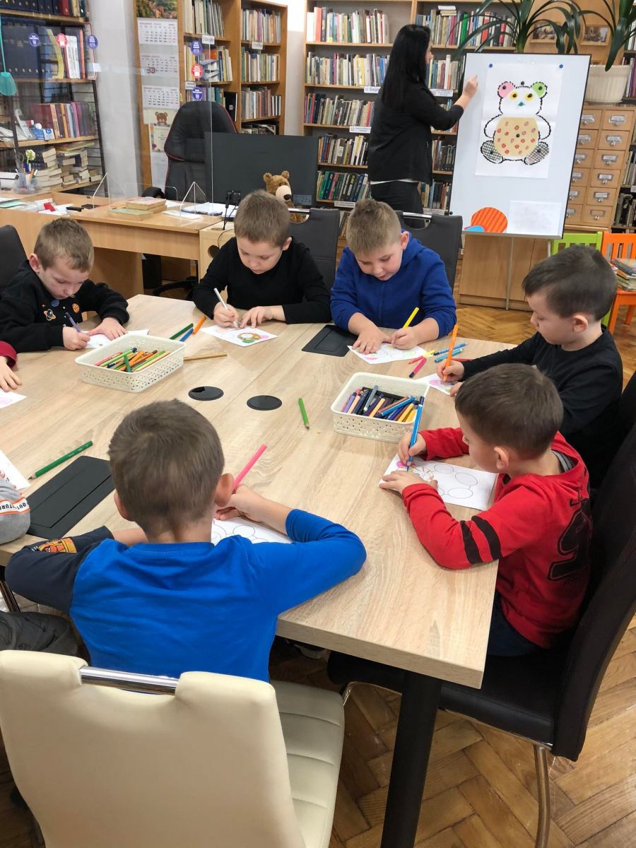 Dzieci z przedszkola kolorują kredkami na stołach w bibliotece rysunek pluszowego misia.
