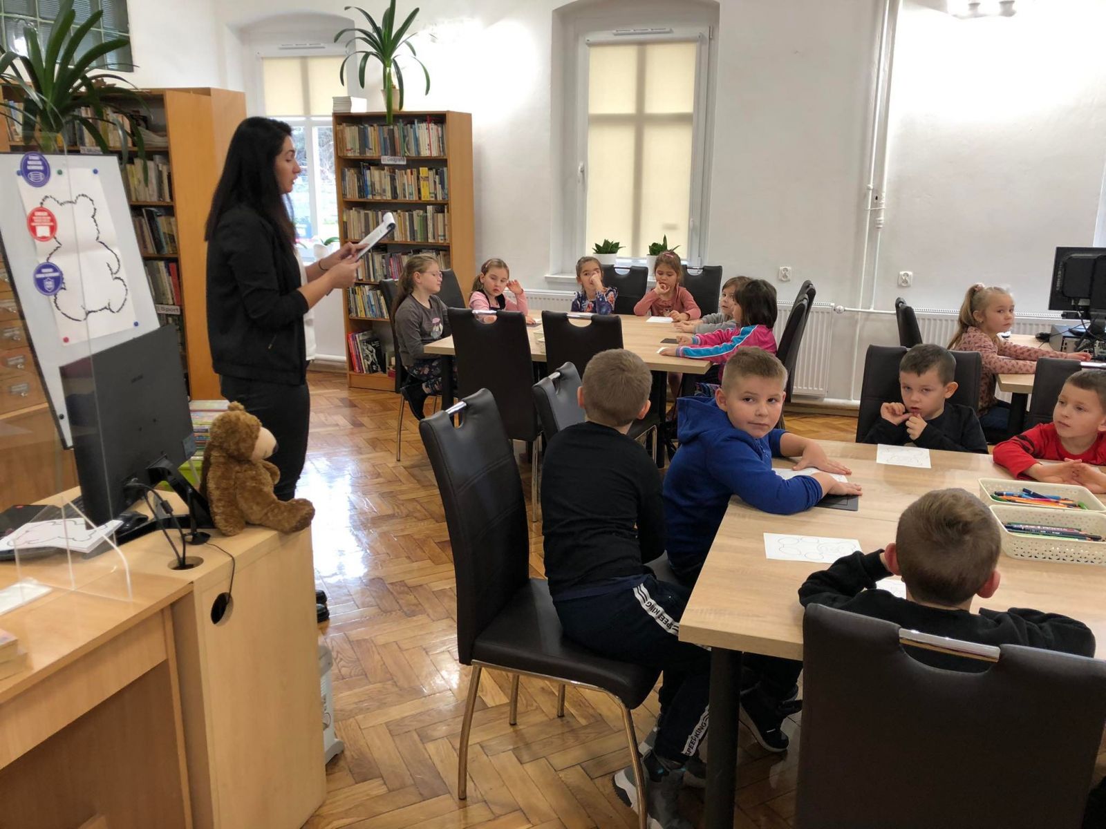 Dzieci z przedszkola w Gromadce siedzą przy stole i słuchają pani bibliotekarki.